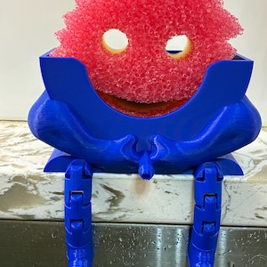 Puede incluir: Un divertido soporte azul para esponja con patas articuladas, diseñado para parecerse a un personaje de dibujos animados. El soporte contiene una esponja roja texturizada con recortes en forma de ojos. Diseño lúdico y funcional.