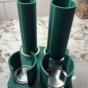 May include: A green 3D-printed kitchen organizer with two tall cylindrical holders and two smaller holders. The holders are designed to hold jars or containers, and are mounted on a rectangular base.