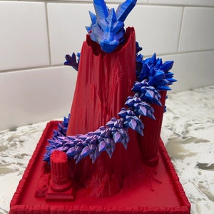Puede incluir: Escultura impresa en 3D de un dragón emergiendo de una montaña roja. La cabeza del dragón y los cristales alrededor de la montaña son azules. La base es roja con bordes decorativos. Una pieza de arte de fantasía única.