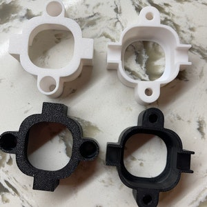 May include: Four 3D-printed plastic objects. Two are white, and two are black. Each has a square-like shape with a circular opening in the center and two circular protrusions. The objects are designed with a textured surface.