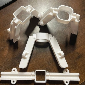 May include: White 3D-printed plastic components, including articulated arms and a rectangular base with mounting holes. The parts are arranged on a wooden surface, suggesting a mechanical or engineering project.