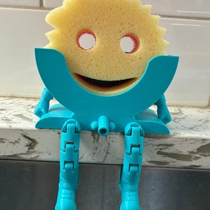 May include: A novelty sponge holder featuring a yellow sponge with eye and mouth cutouts, held by a turquoise plastic frame. The frame has articulated legs, designed to sit on a countertop. A fun and functional kitchen accessory.