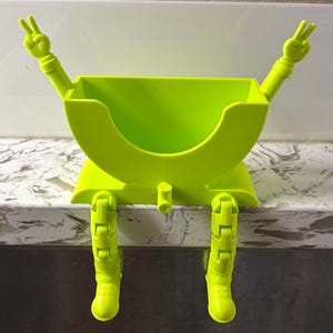 Könnte beinhalten: Ein Schreibtisch-Organizer aus hellgrünem Kunststoff in Form einer sitzenden Figur mit herabhängenden, beweglichen Beinen. Die Figur hat die Arme zum Friedenszeichen erhoben und verfügt über ein offenes Fach zur Aufbewahrung.