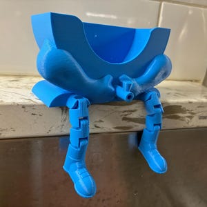 Peut inclure: Un support bleu vif imprimé en 3D doté d'une partie supérieure creuse en forme de bol et de deux jambes articulées pendantes avec des bottes moulées. L'objet est conçu pour être posé sur un rebord, les jambes pendant par-dessus le bord.