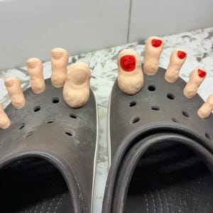 May include: A pair of dark gray Crocs shoes with sculpted toes attached. The toes are painted a flesh tone, with red nail polish on some. The shoes have a white trim around the sole.