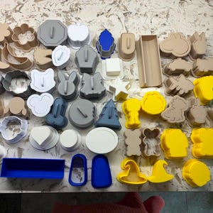 Może przedstawiać: Duża kolekcja plastikowych foremek do ciastek o różnych kształtach i rozmiarach, w tym zwierzęta, gwiazdy, serca i figury geometryczne. Foremki występują w kolorach białym, szarym, beżowym, żółtym i niebieskim, ułożone w rozproszonym układzie.
