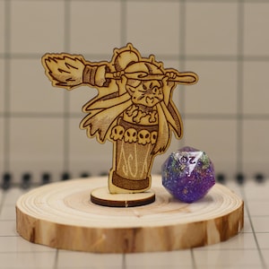 May include: A wooden laser-cut figurine of a witch with a broom, standing on a small wooden base, next to a multi-colored, 20-sided die. The witch figurine has a hat and a skirt with skull designs. The die is purple with flecks of color.