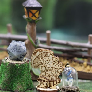 May include: A wooden miniature figurine of a character holding a mushroom and a lantern, standing on a wooden base. A grey and white dice is on a wooden stump with green moss. A glass dome with a blue and white flower inside sits on a wooden base.
