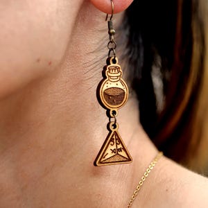 Puede incluir: Pendientes de madera con un diseño de botella de poción y un colgante triangular. Los pendientes son de madera marrón claro y tienen diseños detallados. Cuelgan de un gancho para la oreja de color bronce. El tema es fantasía o magia.