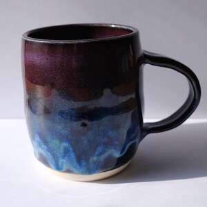 May include: A ceramic mug with a dark purple to blue glaze. The mug has a black handle and a white base. The glaze has a glossy finish. The mug is designed for hot beverages.