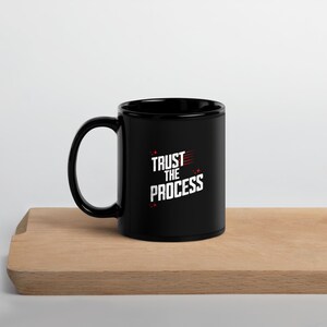 May include: Black ceramic coffee mug with a white inscription that reads "TRUST THE PROCESS" in red lettering.