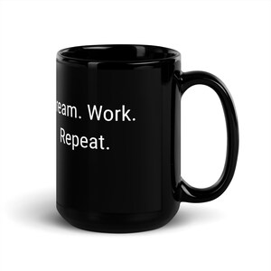 以下が含まれることがあります： 白い文字で「Dream. Work. Repeat.」と書かれた黒いセラミック製のコーヒーカップ