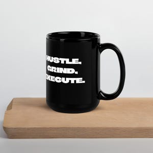 May include: Black ceramic coffee mug with white text that reads "Hustle. Grind. Execute."