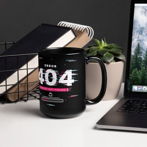 Könnte beinhalten: Schwarze Kaffeetasse mit einer weißen 404-Fehlermeldung und dem Text "Energie nicht gefunden" in Pink. Die Tasse hat auch eine Fortschrittsanzeige mit dem Text "Kaffee wird zubereitet".