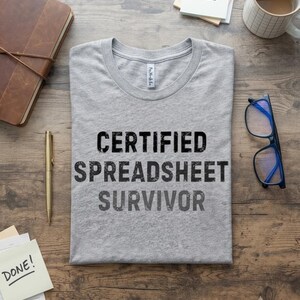 May include: A heather gray short-sleeve t-shirt featuring the black distressed text "CERTIFIED SPREADSHEET SURVIVOR" printed on the front. The shirt has a crew neckline and a standard fit, displayed flat on a wooden surface with office accessories.