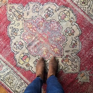 May include: A pair of brown leather ankle boots with pointed toes are worn on a red and cream patterned rug. The rug has a central medallion design.
