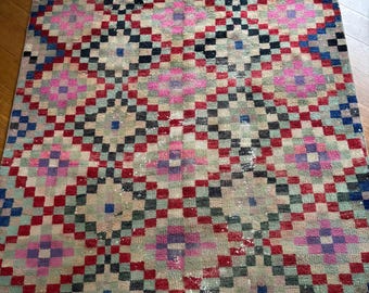 May include: A rectangular rug with a geometric pattern. The rug features a repeating diamond pattern in shades of pink, red, black, blue, and green on a beige background. The rug appears to be made of woven fabric.