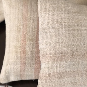 May include: Two off-white throw pillows with a subtle pink stripe pattern. The pillows are made of a woven fabric with a textured surface.