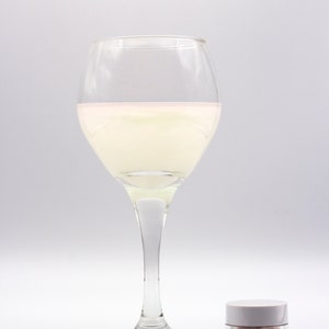 May include: A glass of white liquid with a pink hue in a clear wine glass. A small white jar of edible iridescent glitter is in the foreground. The jar has a black label that reads "Edible Glitter Iridescent NET WT. 0.14 oz (4g)"