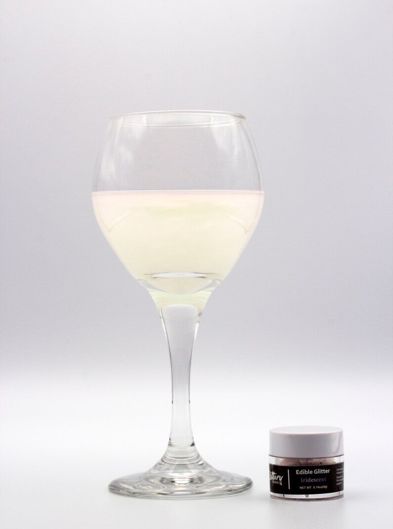 May include: A glass of white liquid with a pink hue in a clear wine glass. A small white jar of edible iridescent glitter is in the foreground. The jar has a black label that reads "Edible Glitter Iridescent NET WT. 0.14 oz (4g)"