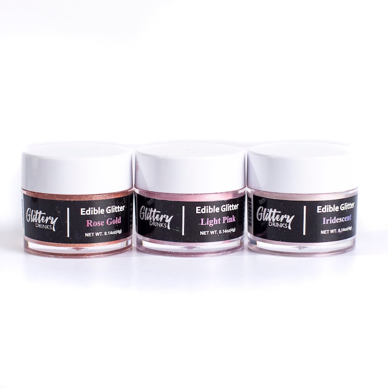 May include: Three small jars of edible glitter in rose gold, light pink, and iridescent colors. Each jar has a black label with the text "Glittery Drinks" and "Edible Glitter" followed by the color of the glitter. The jars are on a white surface.