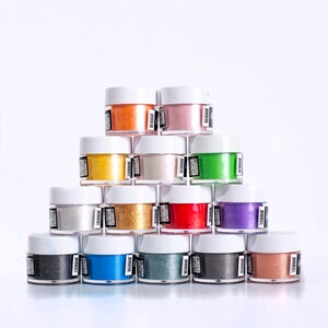 May include: A collection of 14 small jars of colorful glitter, arranged in a pyramid shape. The jars are white with black labels and contain various colors of glitter, including gold, silver, red, blue, green, and purple.