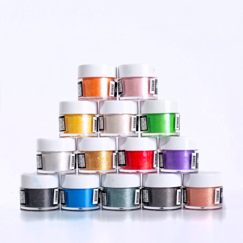 May include: A collection of 14 small jars of colorful glitter, arranged in a pyramid shape. The jars are white with black labels and contain various colors of glitter, including gold, silver, red, blue, green, and purple.