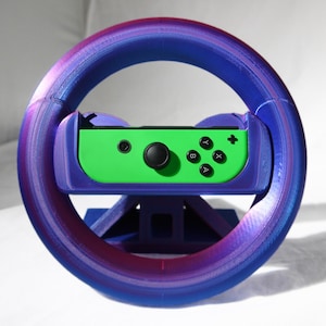 May include: A 3D-printed steering wheel in shades of blue and purple, designed for a gaming console controller. The controller is green with black buttons and directional controls. The wheel is supported by a blue base.