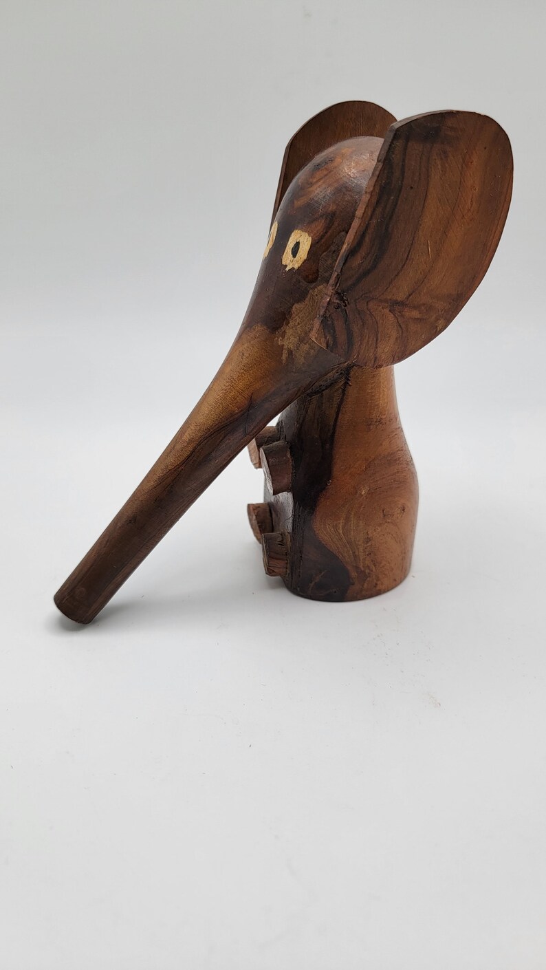 May include: A wooden elephant figurine with a long trunk and large ears. The elephant is carved from dark brown wood and has two small yellow circles for eyes.