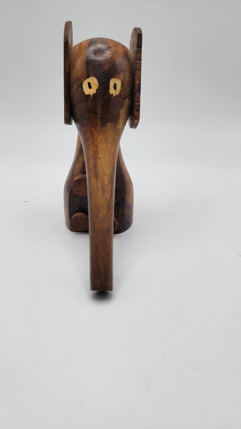 May include: A wooden elephant figurine with a brown and dark brown wood grain pattern. The elephant has large ears and a long trunk. The eyes are inlaid with yellow wood.