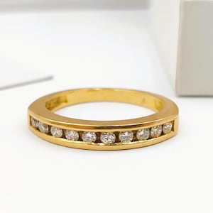 Peut inclure: Une bague en or avec un sertissage en canal, ornée d'une rangée de petites pierres précieuses claires. La bague est posée sur un fond blanc, avec une boîte blanche partiellement visible et un morceau de papier.