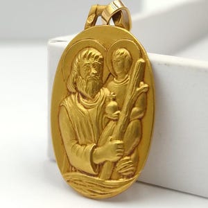Op de afbeelding: Goudkleurige ovale hanger met een reliëf van twee figuren, waarschijnlijk religieus. De hanger heeft een gedetailleerd ontwerp met zichtbare gezichtskenmerken en kleding. Hij hangt aan een kleine lus aan de bovenkant.