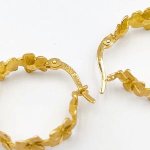 May include: Close-up of two gold hoop earrings. Each earring features a floral design around the hoop. The earrings are open, with the clasp mechanism visible. The background is a solid white.