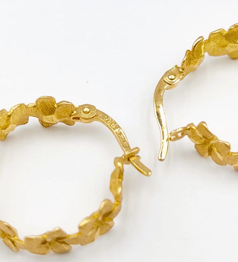 May include: Close-up of two gold hoop earrings. Each earring features a floral design around the hoop. The earrings are open, with the clasp mechanism visible. The background is a solid white.