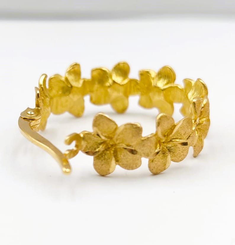 May include: A gold-colored hoop earring featuring a floral design. The earring is adorned with multiple flower-shaped embellishments, creating a continuous pattern around the hoop. The metal has a polished finish, reflecting light.