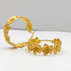 May include: A pair of gold-colored hoop earrings. Each earring is adorned with a series of small, flower-shaped designs. The earrings are set against a white background, highlighting their intricate floral details.