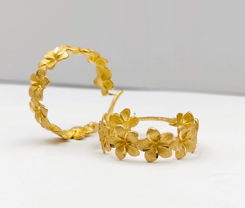 May include: A pair of gold-colored hoop earrings. Each earring is adorned with a series of small, flower-shaped designs. The earrings are set against a white background, highlighting their intricate floral details.