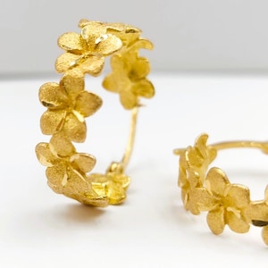 May include: Gold-colored hoop earrings with a floral design. Each earring features a series of small, textured flowers arranged around the hoop. The earrings have a matte finish and a delicate appearance.