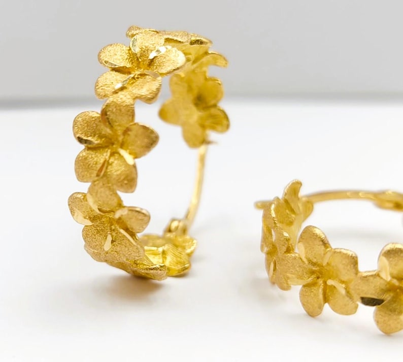 May include: Gold-colored hoop earrings with a floral design. Each earring features a series of small, textured flowers arranged around the hoop. The earrings have a matte finish and a delicate appearance.
