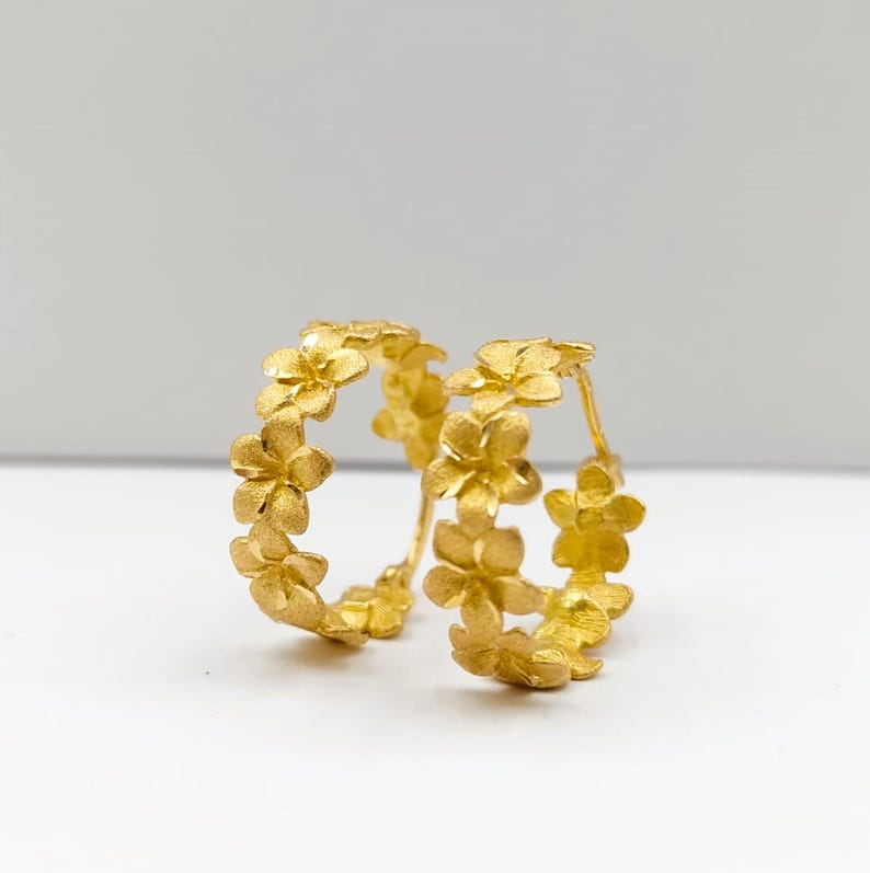 May include: A pair of gold-colored hoop earrings. Each earring is adorned with a series of small, textured flower shapes, creating a floral design. The earrings are set against a white background, highlighting their intricate details.
