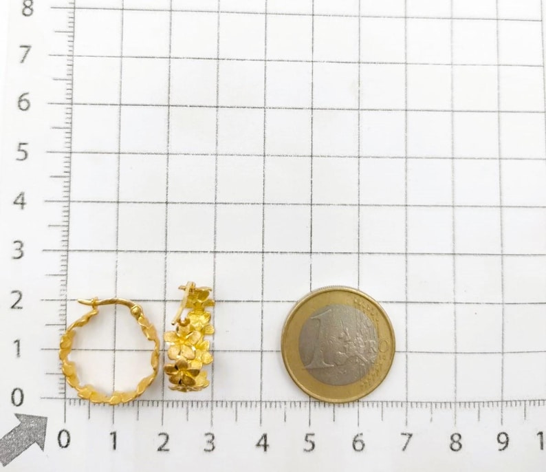 May include: Two gold-colored hoop earrings with a floral design. The earrings are next to a one Euro coin. The earrings are approximately 1 inch in diameter.