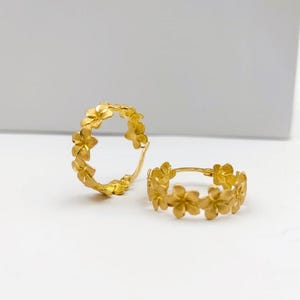 May include: Two gold-colored hoop earrings. Each earring is adorned with a floral design, creating a continuous pattern around the hoop. The earrings are displayed on a white surface.