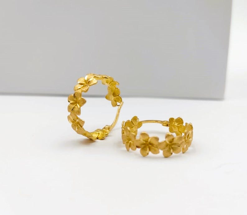 May include: Two gold-colored hoop earrings. Each earring is adorned with a floral design, creating a continuous pattern around the hoop. The earrings are displayed on a white surface.