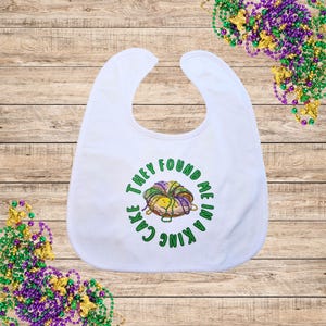 May include: A white baby bib featuring a circular graphic of a king cake decorated with purple, green, and gold beads. The text surrounding the cake reads, "THEY FOUND ME IN A KING CAKE" in green capital letters. The bib has a simple rounded neckline.