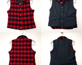 Könnte beinhalten: Wende-Weste mit einem schwarz-roten Büffelkaro-Muster auf der einen Seite und einem einfarbigen schwarzen Design auf der anderen Seite. Die Weste hat einen Kragen, einen Knopfverschluss und zwei Vordertaschen. Sie hängt an einem weißen Kleiderbügel.