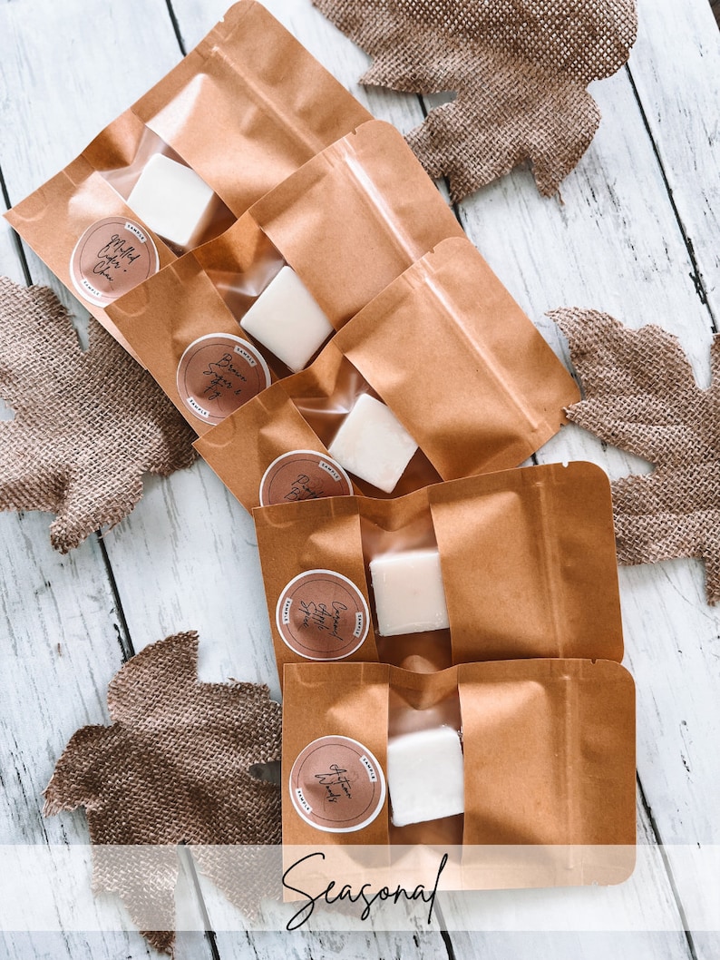 May include: Five brown paper bags containing white wax melts. The wax melts are labelled with different scents: "Mulled Cider", "Brown Sugar & Spice", "Cinnamon Apple Spice", "Autumn Winds", and "Hot Cocoa". The bags are on a white wooden surface with hessian leaves.