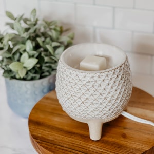 May include: White ceramic wax warmer with a geometric pattern and three legs. A white wax melt is placed in the warmer. The warmer is sitting on a wooden base with black metal legs.