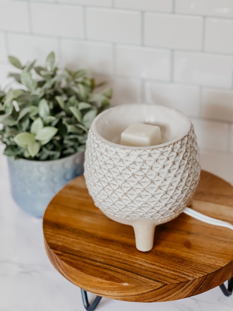 May include: White ceramic wax warmer with a geometric pattern and three legs. A white wax melt is placed in the warmer. The warmer is sitting on a wooden base with black metal legs.