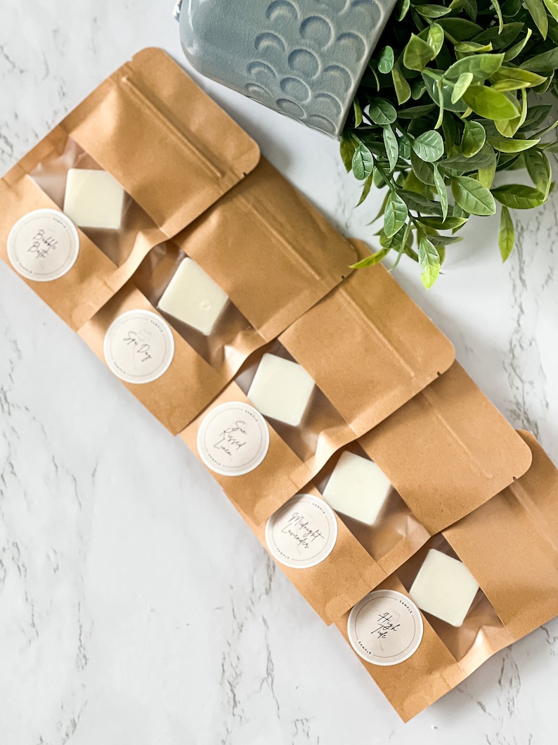 May include: Six brown paper bags with clear plastic windows containing small white wax melts. Each bag has a round label with a different scent name: "Bubble Bath", "Spa Day", "Sweet Cream", "Midnight Lavender", "Hazelnut Coffee", and "Apple Pie".