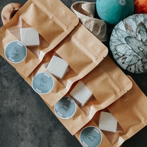 May include: Four brown paper bags containing white wax melts. Each bag features a round sticker with a different scent name: "Pale Sand", "Black Sea", "Smokin' Jacket", and "Sweet Tobacco".